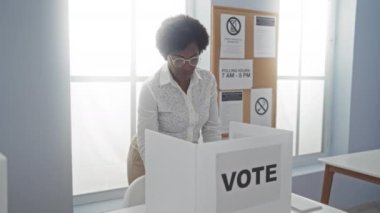 Bir Amerikan oy kullanma merkezinde arka planda ABD bayrağı ve bilgilendirme afişleriyle oy veren bir kadın gururla 