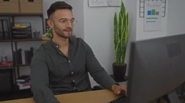 Genç, İspanyol, ofis ortamında gözlüklü, koyu renk gömlekli bilgisayar, raflar ve arka planda bitki olan bir masada çalışan ve duygularını ifade eden bir adam..
