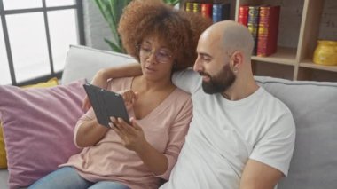 Irklar arası çift rahat bir oturma odasında tabletle kanepede dinleniyor.