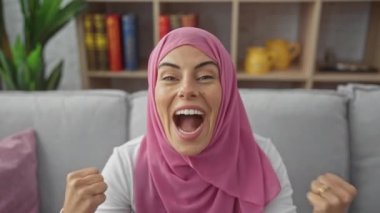 Evde, tesettürlü neşeli genç bir İspanyol kadın zafer kutlamasını kapalı bir yerde kutluyor, kolları koltukta heyecanlı bir zaferle kalkıyor. Gururlu, başarılı ve neşeyle dolu..