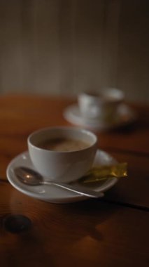 Ön planda beyaz bir kahve fincanı, kaşık ve şeker paketiyle loş bir kafede, ahşap bir masada.