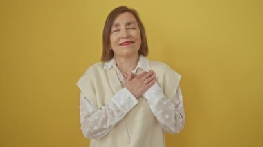 Minnettar orta yaşlı İspanyol kadın, göğsü kapalı, elleri kapalı, yüzünde iç açıcı bir jestle gülümserken sağlık ve mutluluk saçan, izole edilmiş sarı arka planda duruyor..