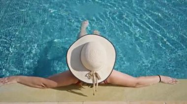Geniş kenarlı bir şapkada gülümseyen bir kadın güneşli bir turkuaz yüzme havuzunun yanında uzanır.