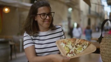 Genç bir kadın İtalya 'da bir açık hava restoranının terasında pizza yemekten hoşlanıyor. Arka planda insanların olduğu hareketli bir sokak sahnesi sergiliyor..