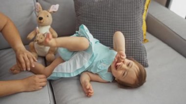 Oturma odasındaki kanepede yeni yürümeye başlamış bir kızla oynayan bir kadın aile sevgisini ve içeride eğlenceli anları arka planda bir oyuncakla sergiliyor.