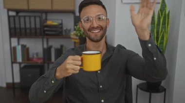 Kitaplık ve bitkilerle dolu bir ofis ortamında elinde sarı bir kupayla gülümseyen ve kameraya el sallayan genç bir adam.