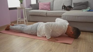 Orta yaşlı İspanyol bir adam evdeki pembe bir paspasın üzerinde yoga yapıyor ve rahat bir oturma odasındaki kanepede iki chihuahua çalıyor..