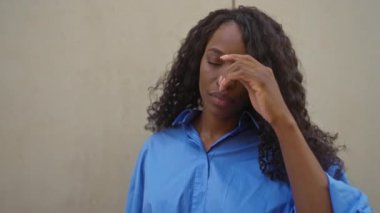 Mavi tişörtlü, kıvırcık saçlı, beyaz bir duvar arkasında poz veren güzel, Afro-Amerikalı bir kadın..