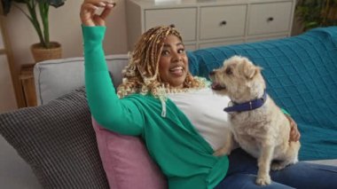 Kapalı alanda örgü ören Afro-Amerikalı genç bir kadın evinin oturma odasındaki kanepede otururken köpeğine seve seve ikram ediyor..