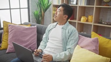 Gözlüklü, düşünceli Asyalı bir adam gri bir koltukta oturuyor ve rahat bir oturma odasında dizüstü bilgisayar kullanıyor.