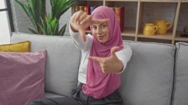 Tesettürlü, neşeli, genç İspanyol bir kadın evinde elleri kanepenin üzerinde bir fotoğraf çerçeveletiyor. Mutlu fotoğrafçılık ve yaratıcılık kavramını somutlaştırıyor.