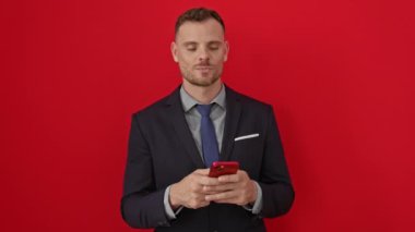 Genç İspanyol takım elbiseli adam komik balık suratı yapıyor çılgın gözleri ve ağzı ile izole edilmiş kırmızı arka planda akıllı telefon kullanıyor.