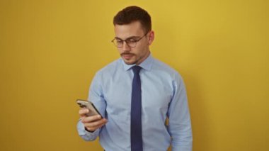Genç İspanyol adam gömlek ve kravat takıyor, akıllı telefon kullanıyor, göz kırpıyor ve izole edilmiş sarı arka planda seksi bir ifadeyle mutlu görünüyor.