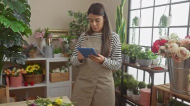 Çizgili gömlek ve önlük giyen genç bir İspanyol kadın çiçeklerle dolu bir evin içinde dururken elinde bir tablet ve sarı bir çiçek tutuyor..