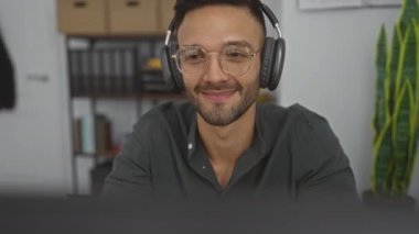 Kapalı ofis ortamında masa başında çalışırken gülümseyen gözlüklü ve kulaklıklı İspanyol adam..