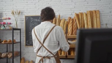 Fırında çalışan bir adam çeşitli ekmek ve hamur işleriyle dolu rafların önünde duruyor, gülümsüyor ve maşa tutuyor, beyaz gömlek ve kahverengi önlük giyiyor.