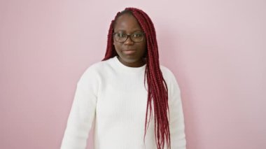 Gözlüklü ve beyaz kazaklı kendinden emin Afrikalı bir kadın pembe bir duvara karşı baş parmak hareketi yapıyor..