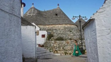 Yeşil elbiseli genç İspanyol kadın, açık mavi gökyüzünün altındaki sembolik Trulli evleri ile İtalya 'nın büyüleyici eski Alberobello kasabasında yürüyor ve Puglia' nın özünü yakalıyor..
