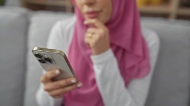 Modern bir evde akıllı telefon tutan tesettürlü düşünceli bir kadın..