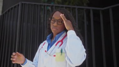 Güzel bir Afro-Amerikalı kadın hemşire şehir ortamında bir hastanenin önünde duruyor, kafasını sıkmış, stresli ya da yorgun görünüyor, boynunda steteskopla..