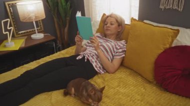 Sarışın bir kadın, rahat bir yatak odasında kitap okuyor. Yanında da ev dekoru ve sıcak ışıklandırmayla çevrili bir köpek yatıyor..