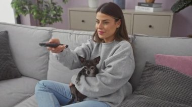 Bir kadın sıcak bir oturma odasında uzaktan kumandayla kanepede bir chihuahua 'ya sarılıyor..