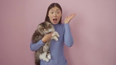 Genç Çinli kadın ve kedi açık ağızla bir arada duruyorlar, şaşırmış ve korkmuş, pembe arka plandaki yüzlere güvenmiyorlar.