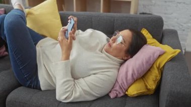 Rahat bir kadın rahat bir oturma odasında akıllı telefonunu kullanarak koltuğa yaslanır.