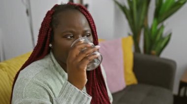 Örgü ören Afrikalı bir kadın evde dinlenirken su içiyor. Etrafı rahatlatıcı oda dekoruyla çevrili..