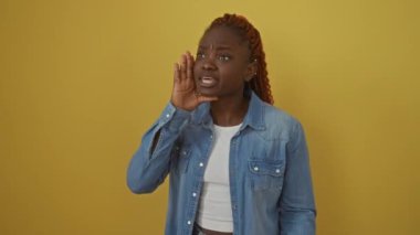 Kot pantolonlu Afrikalı bir kadın, ayakta duruyor ve elleri ağızlarının üstünde bağırıyor, bağırıyor, bağırıyor, sarı bir arka planda.