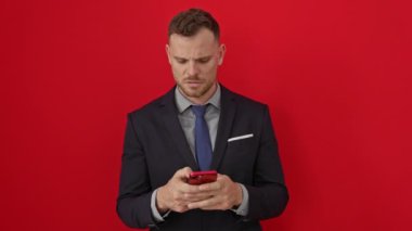 Yüzünde şüpheci bir ifade olan İspanyol takım elbiseli genç adam izole edilmiş kırmızı arka plan üzerinde akıllı telefon kullanarak alaycı bir şekilde şok oldu.