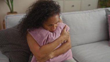 Oturma odasında kanepede yastığa sarılan kadın, günlük giysiler giyiyor, hüzün, yalnızlık ve ev ortamında tefekkür duygularını paylaşıyor.