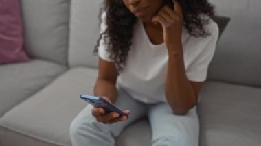 Genç Afro-Amerikalı kıvırcık saçlı kadın oturma odasında oturuyor, akıllı telefon kullanıyor ve düşünüyor..