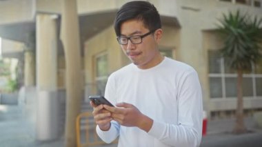 Neşeli Asyalı gözlüklü bir adam güneşli bir şehir sokağında akıllı telefon kullanıyor, günlük ve bağlantılı bir hayat sürüyor..