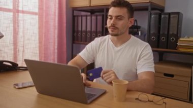 Laptopta çalışan yakışıklı İspanyol bir adam. Elinde klasörleri ve rafları olan bir ofis odasında kredi kartı tutuyor..