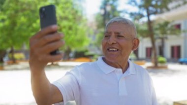 İspanyol beyaz saçlı adam şehir parkında akıllı telefonuyla bir video görüşmesi yapıyor. Şehir zemininde ve yeşillikte gülümsüyor..