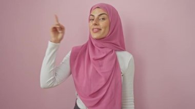 Tesettürlü neşeli genç İspanyol kadın kendine güvenen bir numarayı işaret ediyor, parlak mutlu bir yüz izole edilmiş pembe arkaplan üzerine fikirlerle dolu.