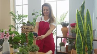 Kırmızı önlüklü, gülümseyen kadın ev ortamında, kapalı alandaki bitkiler arasında akıllı telefon kullanıyor.