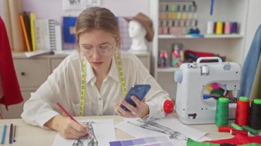 Bir terzinin atölyesinde akıllı telefon kullanan odaklanmış genç bir kadın. Tasarım çizimleri, dikiş makinesi ve kumaşla çevrili..