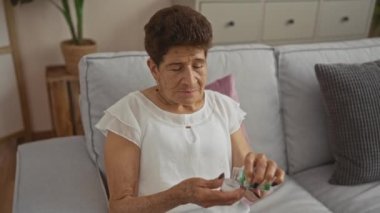 Kısa saçlı yaşlı İspanyol kadın, gri bir kanepede rahat bir oturma odasında oturmuş ilaç şişesini açarken beyaz bir bluz giyiyor..