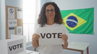 İspanyol orta yaşlı kadın Brezilya seçim okulunda el ve parmak ile gülümserken oy kağıdına mutlulukla işaret ediyor.