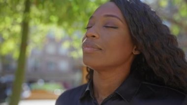 Kıvırcık saçlı güzel bir kadın yeşil ağaçlar ve şehir binalarıyla çevrili şehir parkında dalgın dalgın görünüyor..