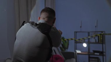 Rozeti olan odaklanmış bir adam, elinde bir el feneri ve silahla, kapalı alanda hafif aydınlatılmış bir suç mahallini dikkatle araştırıyor..