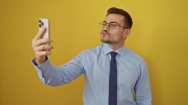 Tişörtlü ve kravatlı genç adam akıllı telefon kullanıyor, kameraya aşk ifadesi ile flört öpücüğü veriyor, izole edilmiş sarı arka planda seksi ve sevimli görünüyor.
