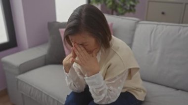 Orta yaşlı üzgün bir kadın elleriyle yüzünü kapatıyor, modern bir oturma odasında kanepede oturuyor.