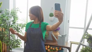 Önlüklü gülümseyen kadın elinde telefonla, kapalı mekan bitkilerinin arasında görüntülü konuşma yapıyor.