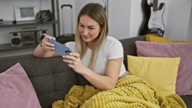 Neşeli genç bir kadın, rahat, modern bir oturma odasında akıllı telefonuyla meşgul olurken koltukta rahatlar ve güler..