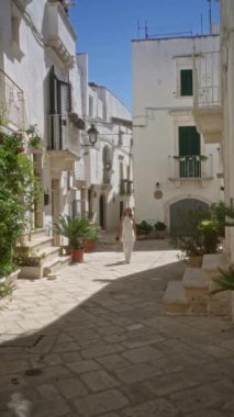 Genç bir İspanyol kadın açık mavi bir gökyüzünün altında beyaz boyanmış binalar ve saksı bitkileriyle çevrili locorotondo, puglia, İtalya 'nın büyüleyici eski caddelerinde yürüyor..