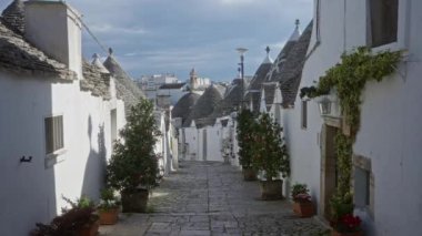 Alberobello, İtalya 'da pitoresk bir caddede taş çatılı geleneksel trulli evler ve parke yolu boyunca yürüyen bir adam, saksı bitkileri ve açık mavi gökyüzü ile çevrili.