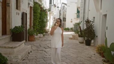 Beyaz elbiseli genç bir İspanyol kadın, beyaz binalar ve yeşilliklerle çevrili Puglia, İtalya 'nın eski locorotondo caddelerinde yürürken dondurma yiyor..